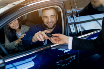 Smiling millennial couple receiving car keys from salesman while sitting inside their newly...