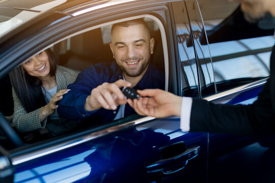 Smiling millennial couple receiving car keys from salesman while sitting inside their newly purchased car in dealership showroom, happy spouses buying modern vehicle in auto salon, closeup