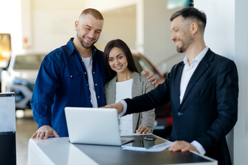 Caucasian millennial couple consulting with salesman about car purchase using laptop in dealership showroom, happy young spouses checking options before buying vehicle in modern auto center