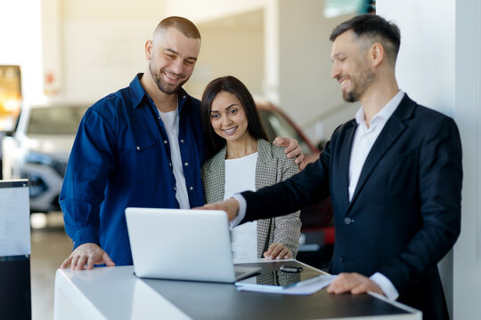 Caucasian millennial couple consulting with salesman about car purchase using laptop in dealership showroom, happy young spouses checking options before buying vehicle in modern auto center