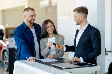 Happy caucasian millennial couple receiving car keys from salesman after signing contract in...