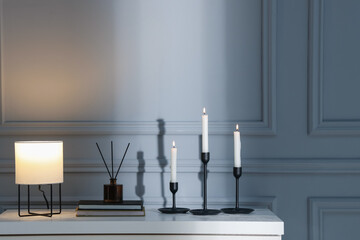 Stylish candlesticks with burning candles, books and other decor on white table near gray wall...