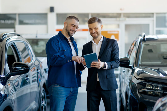Caucasian millennial salesman showing digital tablet to a male customer, manager in suit and buyer standing between cars in modern dealership showroom, discussing auto characteristics