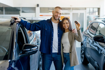Joyful couple showing car keys together, smiling in modern dealership showroom, happy young man and woman excited about their purchase, posing in auto salon, looking at camera