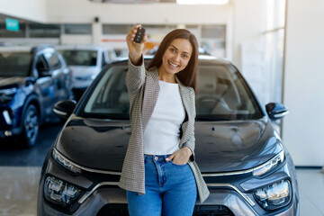 Happy young woman showing car keys at camera while standing in auto showroom, millennial smiling...