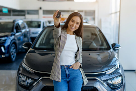Happy young woman showing car keys at camera while standing in auto showroom, millennial smiling female celebrating purchasing new vehicle at dealership center , copy space
