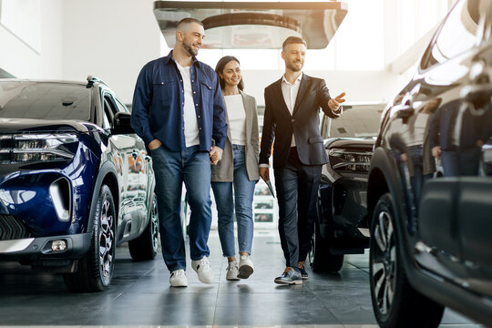 Smiling car salesman showing vehicles to a cheerful couple, buyers family and salon manager walking together inside spacious dealership showroom, considering options - Powered by Adobe