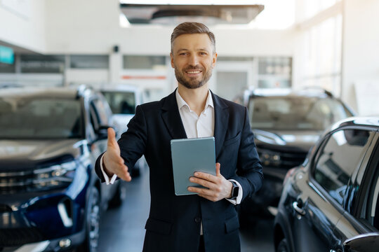 Car salesman holding digital tablet, greeting and extending hand for handshake, smiling warmly while working at the dealership center with modern vehicles in the background, copy space