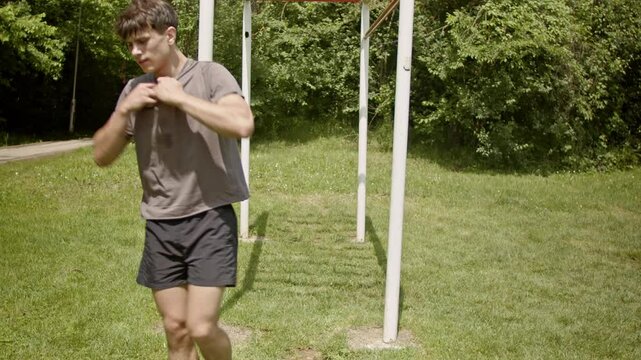 A young man takes off his shirt and does pull-ups on an outdoor fitness structure. He is working out in nature.