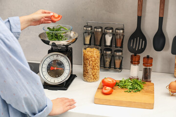 Woman adding tomato into bowl on mechanical kitchen scale at white marble countertop indoors,...