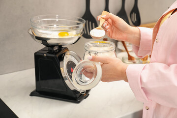 Woman holding spoon with flour near mechanical kitchen scale at white marble countertop indoors,...