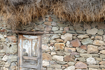 Fragment of a wall of an ancient building made of stones. Wall of a building made of stones and a thatched roof. A roughly made door in a stone wall and with a fragment of a thatched roof.
