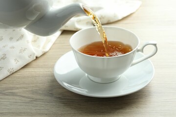 Pouring aromatic tea into cup from teapot at wooden table, closeup