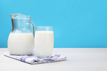 Fresh milk in glass and jug on white wooden table against light blue background. Space for text