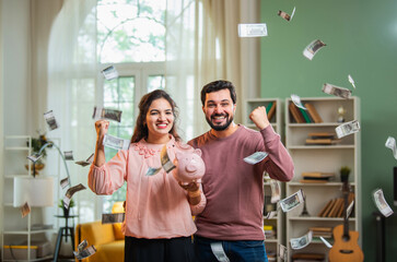 Happy Indian man and woman saving money in piggy bank as currency rain falls in living room