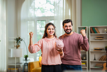 Happy Indian man and woman saving money in piggy bank as currency rain falls in living room