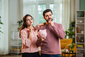Young Indian couple unsure about saving plans while holding piggy bank in living room