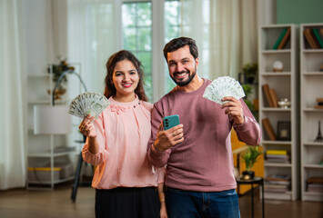 Happy Indian couple with currency note fan and phone, celebrating lottery or business profit