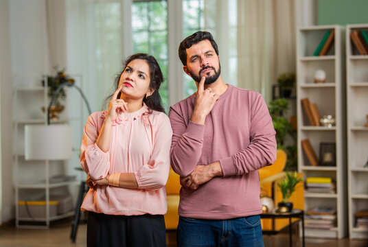 Indian couple standing at home with confused expressions, thinking about decision or problem