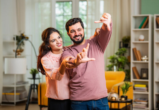 Happy Indian couple imagining house makeover with hand frame in cozy living room - Powered by Adobe