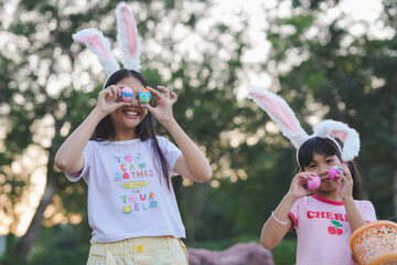 Two girls have fun finding Easter eggs a favorite activity for all kids. The girls wear cute bunny ears on the head holding a basket with Easter eggs in a natural garden with evening light.