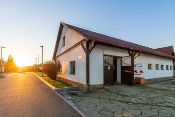 Obraz premium Warm sunlight casts a golden glow on a farmhouse at sunset, with a calm country road leading to the entrance. The peaceful atmosphere highlights simplicity and rural charm.