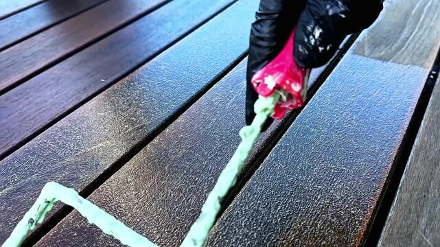 A skilled worker applies wood protection to an outdoor hard wood deck using a roller, ensuring an even application across the surface