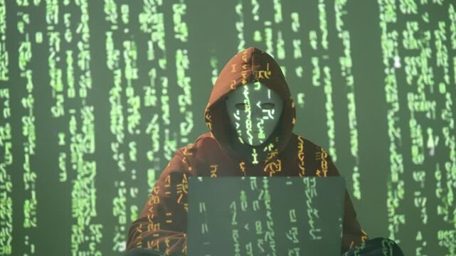 Haker w masce i kapturze pracujący na laptopie w otoczeniu zielonego kodu matrix, cyberprzestępczość i bezpieczeństwo cyfrowe w mrocznym, anonimowym środowisku