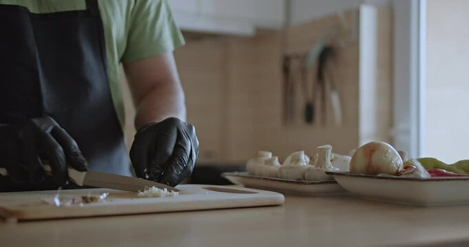 A man wearing black gloves carefully chops garlic on a wooden cutting board, preparing ingredients for a meal.