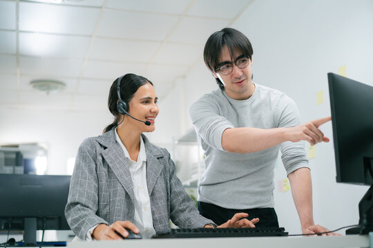 customer service representative wearing headset is seated desk, smiling while working computer. colleague stands beside her, pointing screen, assisting with task call center environment