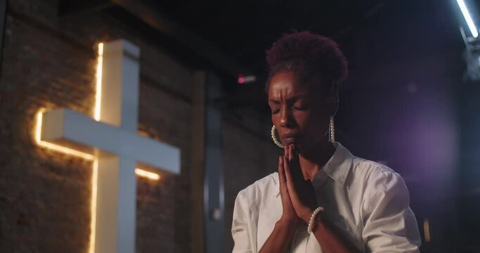 Young Black woman praying in a church, hands clasped, eyes closed in deep contemplation, glowing cross in background, symbolizing faith, devotion, and spirituality