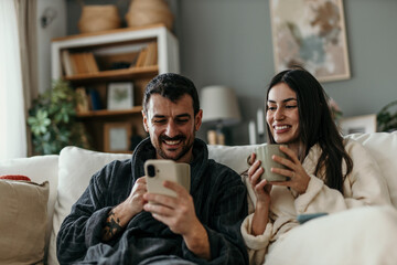 Young smiling couple wearing bathrobes, sitting on sofa and enjoying morning coffee, man using smartphone