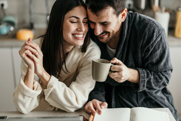 Young loving couple wearing bathrobes, sharing a cup of coffee and smiling while planning their day together in the kitchen
