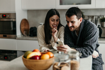 Cheerful man and woman wearing bathrobes, using mobile phone and smiling while having breakfast in modern kitchen