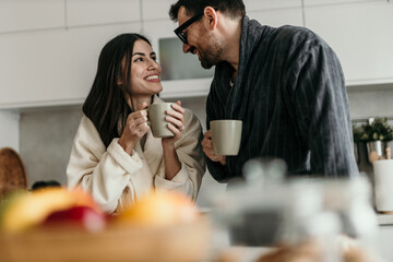 Young smiling couple wearing bathrobes, drinking coffee and looking at each other during breakfast in a modern kitchen