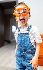 Kid in goggles shows joy indoors. Young child dressed in overalls and protective goggles expresses...