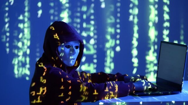 Młody haker w masce i bluzie z kapturem pracuje przy laptopie w świetle cyfrowych kod&oacute;w matrix na niebieskim tle, symbolizując cyberprzestępczość, technologię i zagrożenia online