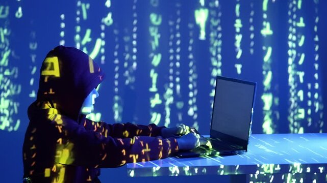 Anonimowy haker w ciemnej bluzie z kapturem pracuje przy laptopie w niebieskim świetle z kodem matrix w tle, symbolizując cyberatak, cyfrowe bezpieczeństwo i hakerską działalność nocą