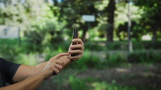Man using smartphone in a park with a green garden background, showcasing technology interaction in a serene outdoor environment.