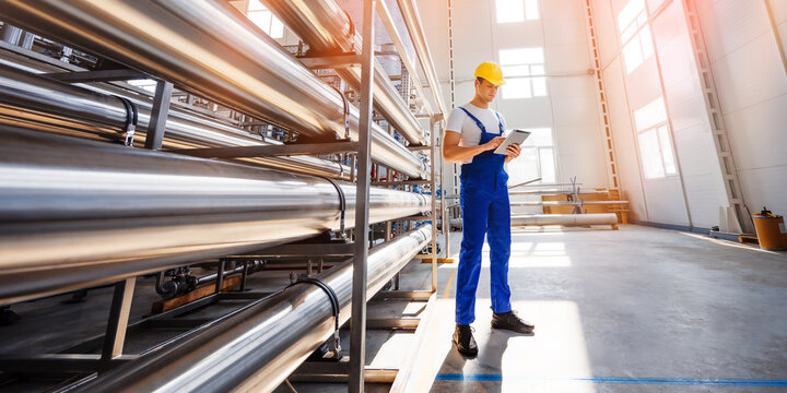 Young male specialist in a yellow hard hat uses tablet PC for controls the technological processes of equipment for industrial water purification. Industrial worker indoors in factory.