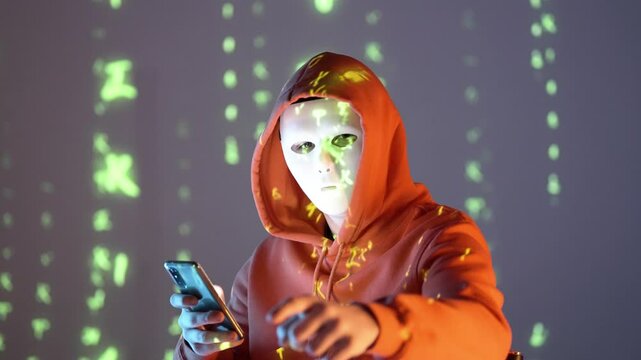 Osoba w masce i kapturze wskazująca w kamerę, z telefonem w ręku, na tle zielonego kodu &ndash; haker, cyberprzestrzeń, dane, technologia