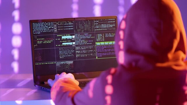 Osoba w kapturze pracująca na laptopie z kodem na ekranie, otoczona cyframi binarnymi &ndash; haker, cyberatak, dane, kodowanie, systemy