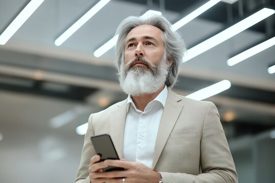 Contemplative older man with beard holding smartphone looking up thoughtfully