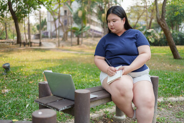 chubby asian woman sits on park bench, focused on her laptop and taking notes in notebook,...