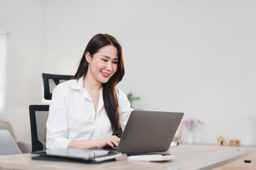Asian businesswoman in white shirt works happily on laptop at modern office desk