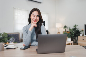 Asian businesswoman working on laptop in modern office, smiling confidently while multitasking with...