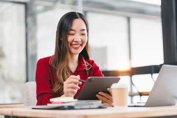 Obraz premium Asian businesswoman in red blouse smiles while working on tablet at modern office desk with laptop and coffee