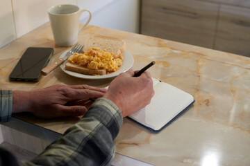 Man writing notes in notebook, eating scrambled eggs on toast, coffee beside smartphone at kitchen counter during morning routine