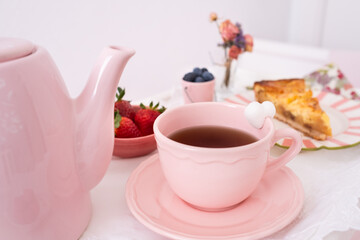 Detail of a delicious breakfast on a bed in white and pink tones. Natural light enters through the window. There is a teapot, a tray, fruit , a sprig of flowers and an apple pie. Care and love