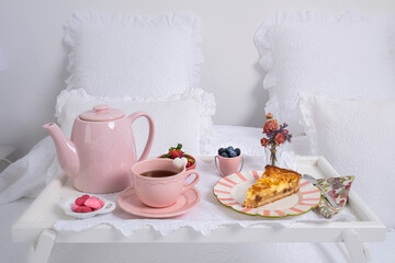 high angle view of a beautiful breakfast on a white bed. Natural light illuminates a teapot, a tray, fruit , a sprig of flowers and an apple pie. Care and love. Birthday love detail or Valentine day